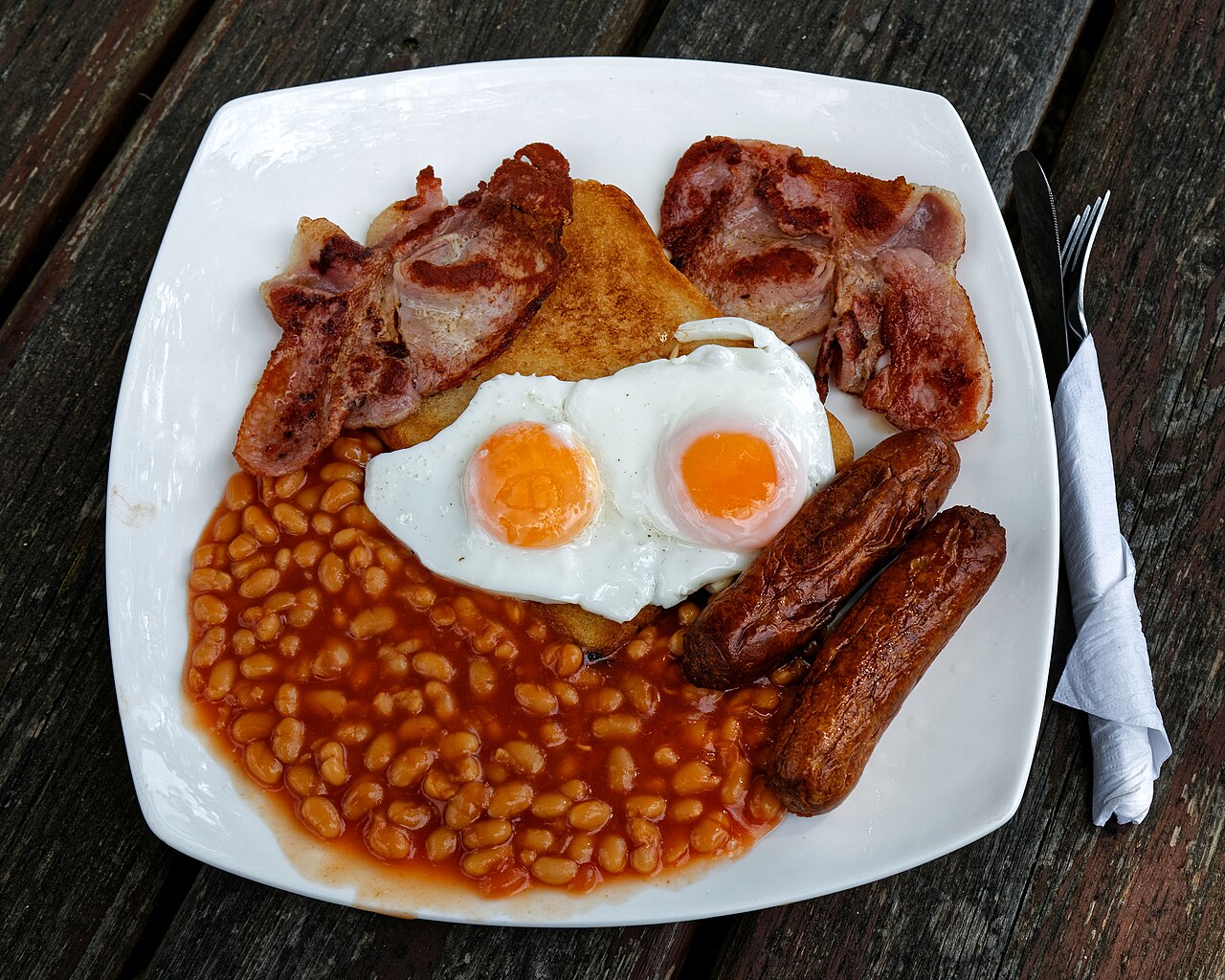 Brunch plate with two eggs, bacon, sausages, beans, and toast on a square white plate
