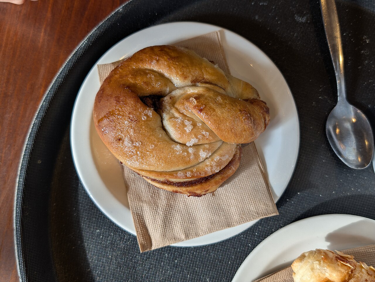 Sugar-crusted knotted cardamom bun on a small plate with a spoon beside