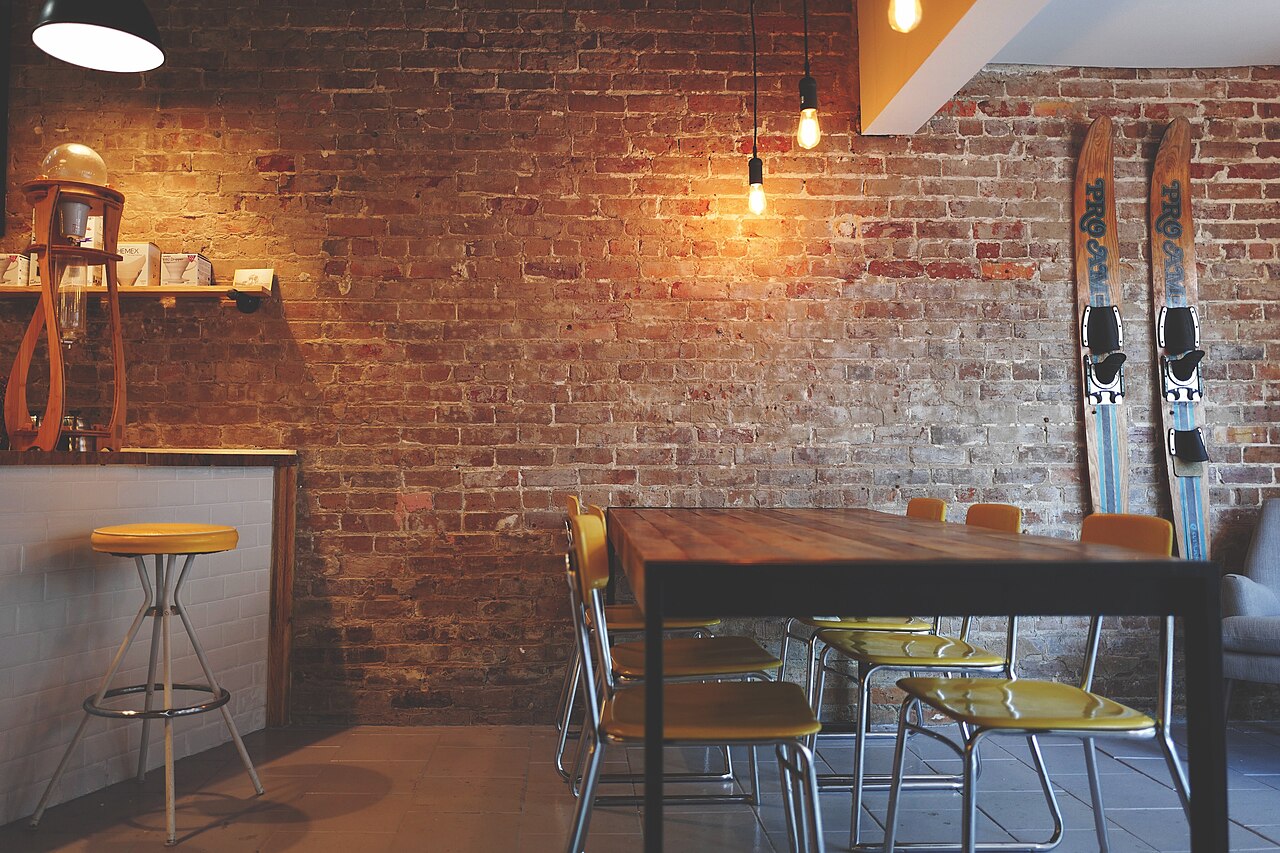 Café interior with brick wall, hanging Edison bulbs, mustard-yellow chairs around a wooden table